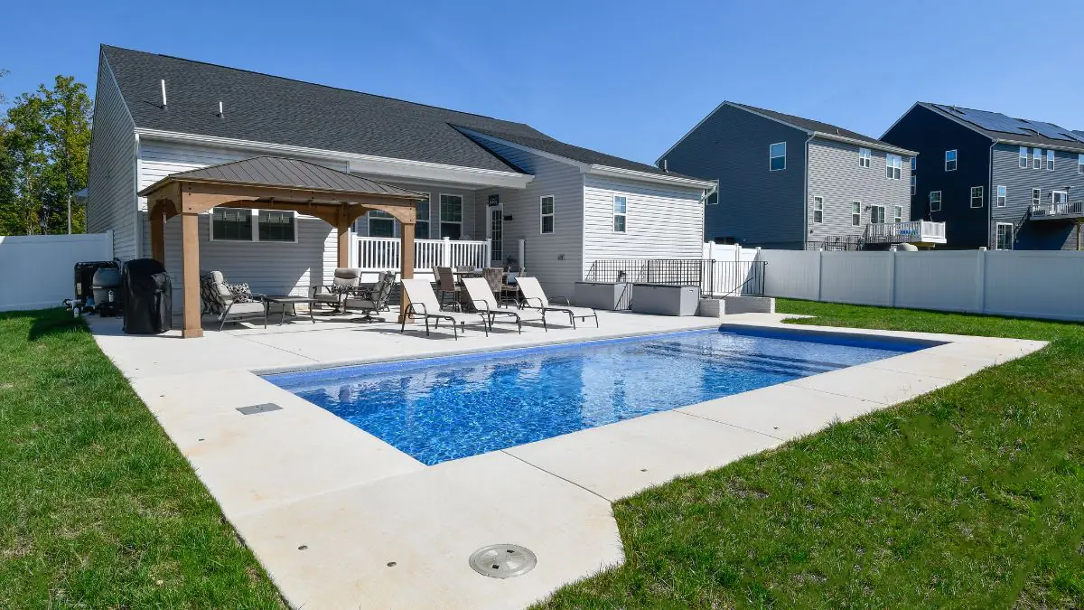 Backyard pool with concrete patio, wood pergola, lounge chairs, and white vinyl fencing