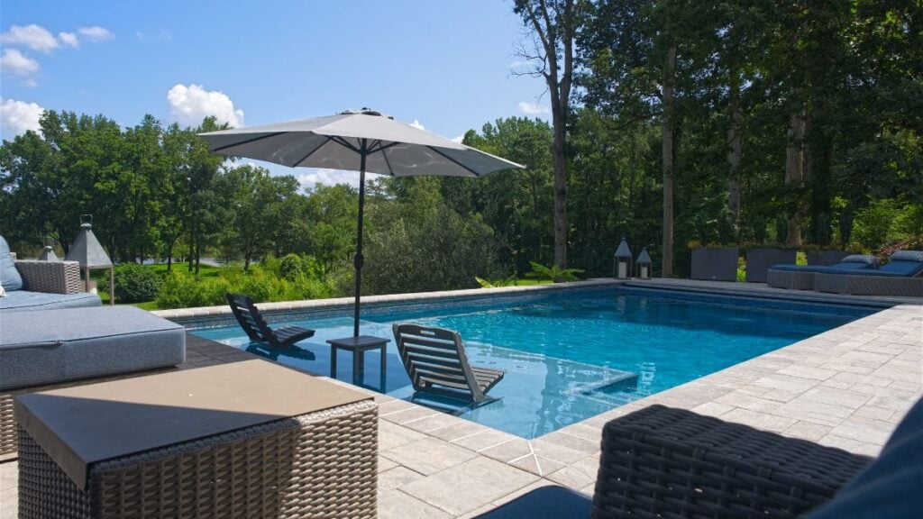 Pool with in-water lounge chairs and umbrella, framed by wicker patio furniture and trees