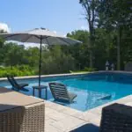 Pool with in-water lounge chairs and umbrella, framed by wicker patio furniture and trees
