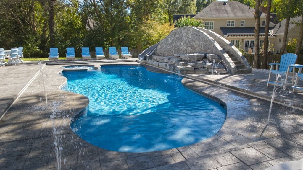Freeform pool with a stone waterslide bridge, deck jets, and blue lounge chairs on a patio