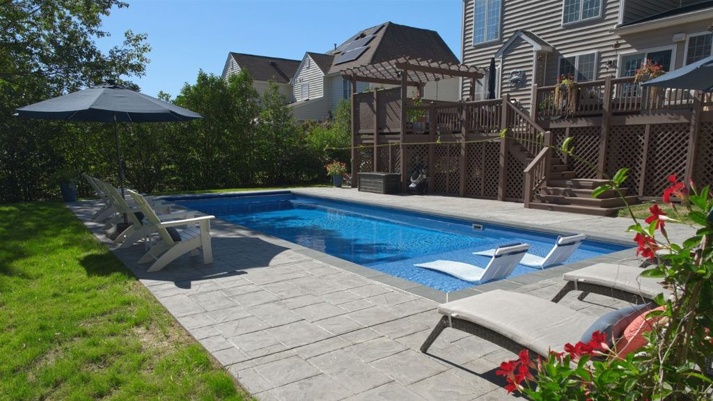 Inground pool with tanning ledge, in-pool loungers, and spacious stone patio beside a home