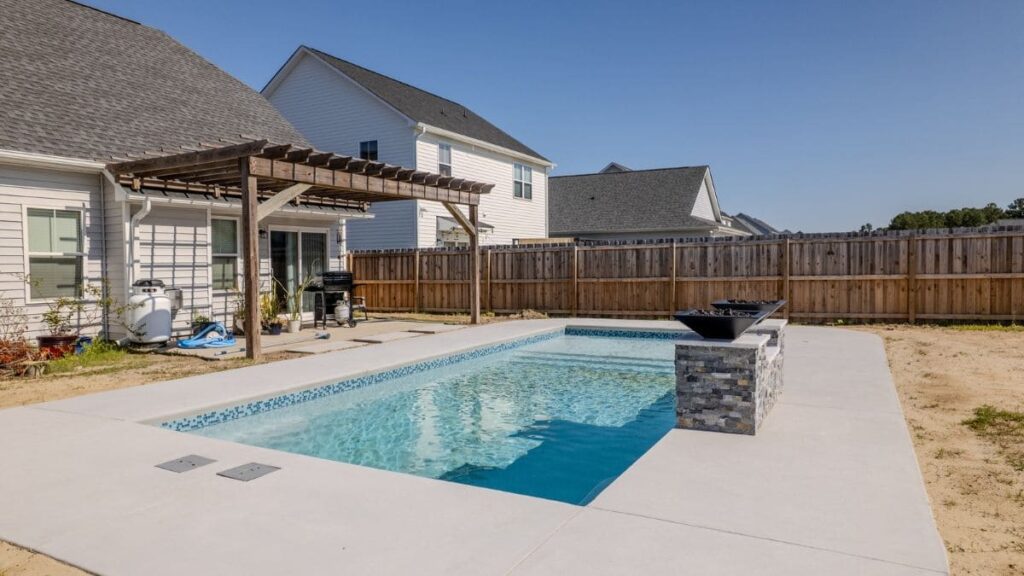 Small rectangular backyard pool with a blue tile border, light concrete deck, stacked stone pillar with fire bowls, and a wood pergola beside the home