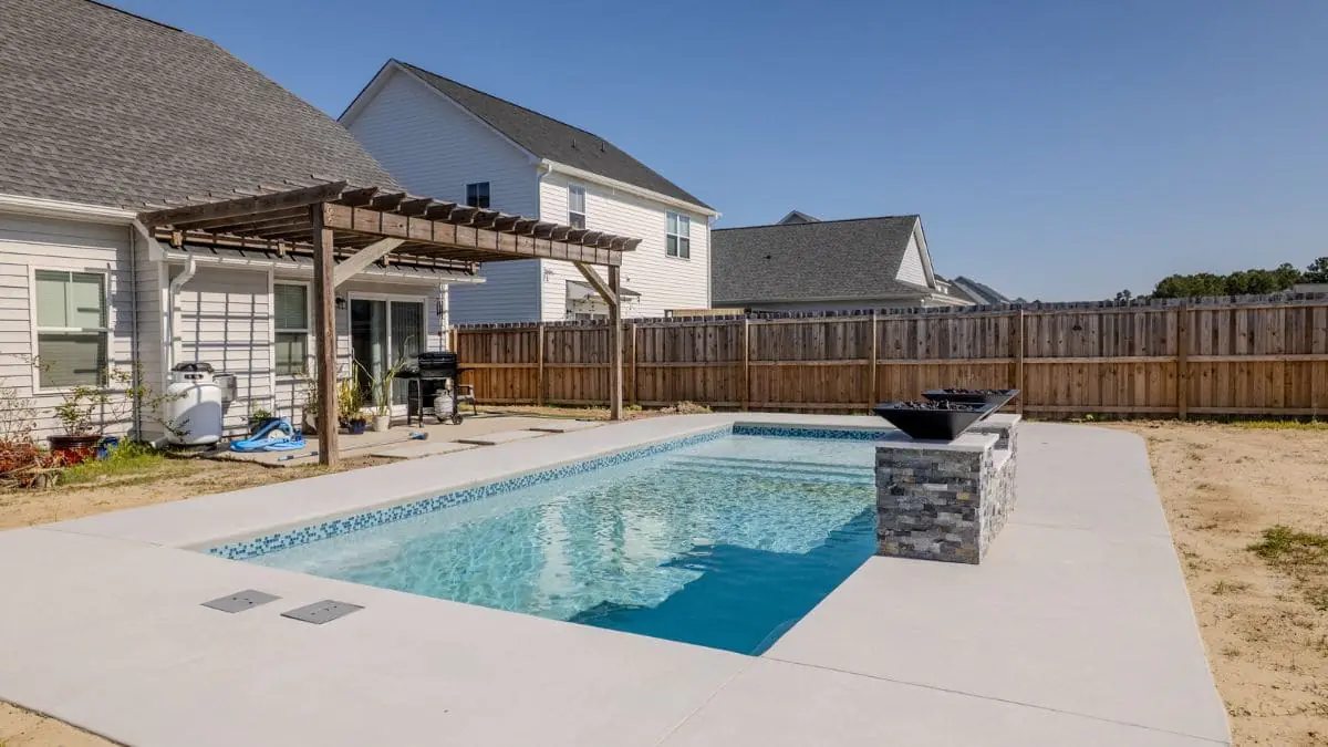 Small rectangular backyard pool with a blue tile border, light concrete deck, stacked stone pillar with fire bowls, and a wood pergola beside the home