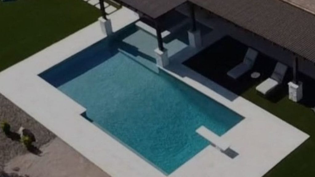 Aerial view of a rectangular backyard pool with a white concrete deck, diving board, covered patio with lounge chairs, and artificial turf