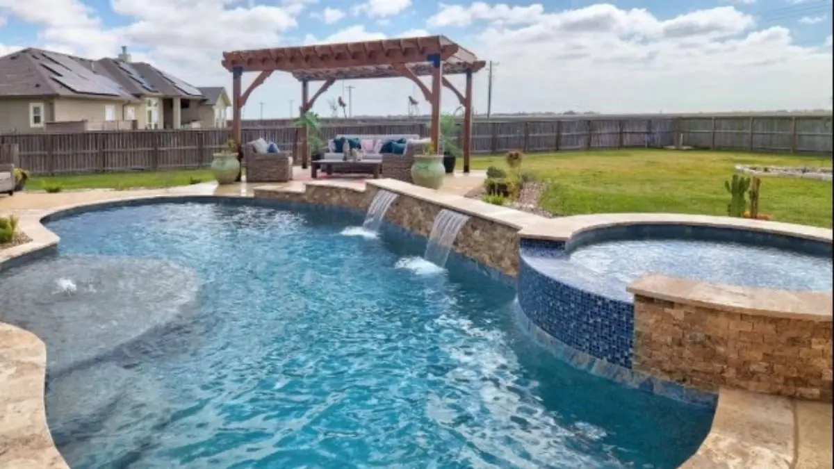 Freeform inground pool with stone waterfall features, raised spa with blue tile, and a pergola seating area in a backyard