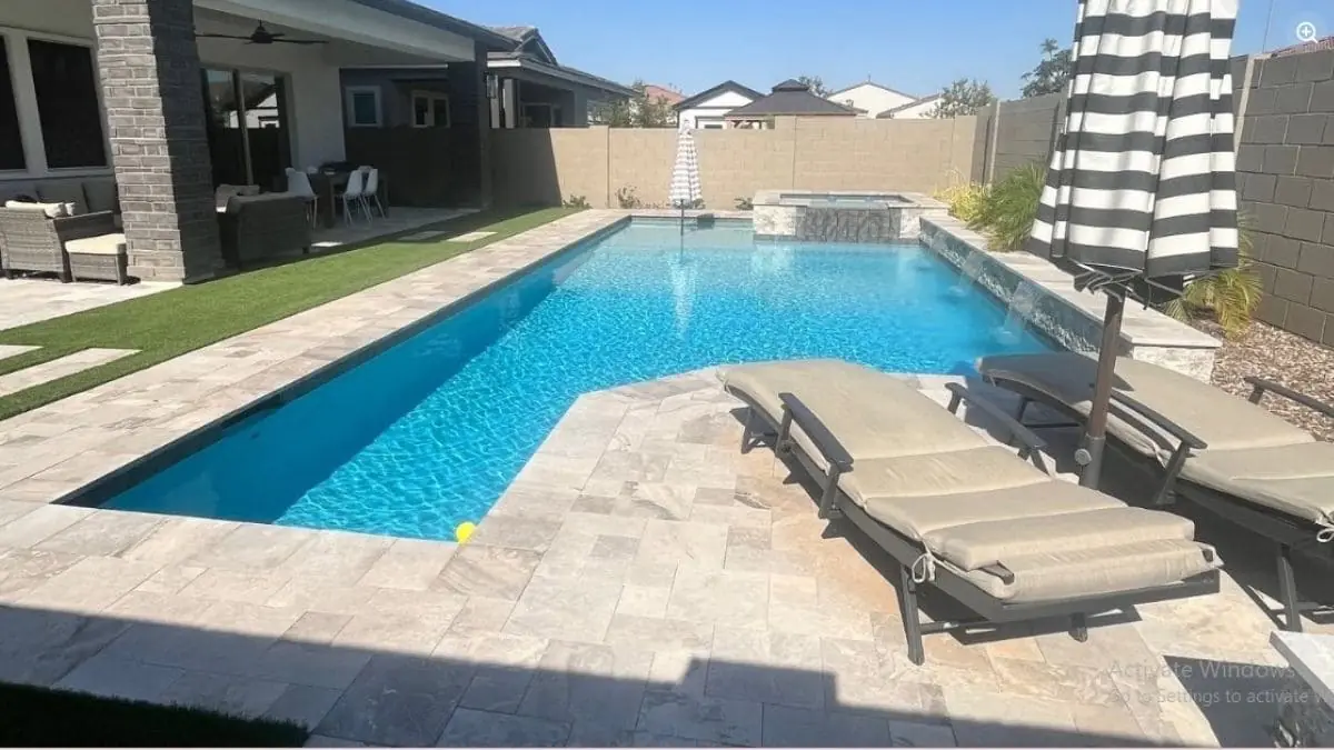 Backyard pool with bright blue water, travertine deck, cushioned lounge chairs, a striped umbrella, and a raised spa with a water feature
