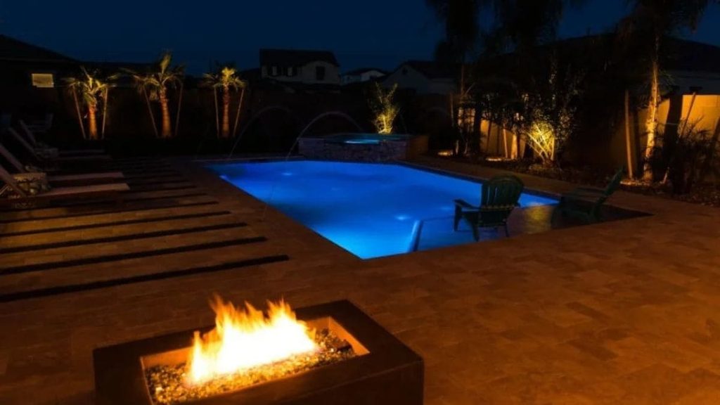 Backyard inground pool lit blue at night with a glowing fire pit, paver patio, lounge chairs, and uplit palm trees