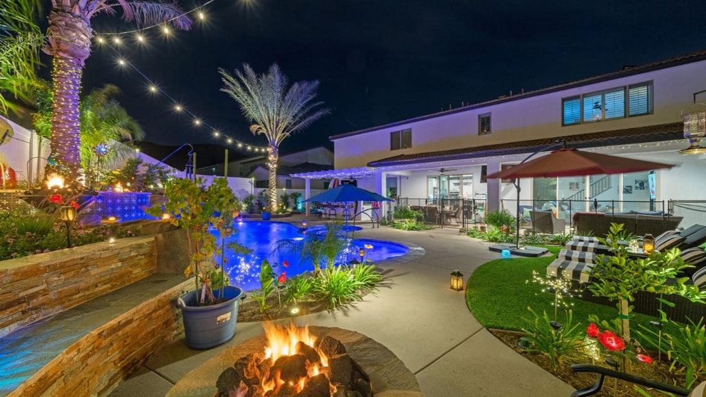 Backyard pool glowing blue at night with a stone fire pit, string lights on palm trees, a raised stacked stone planter, umbrellas, lounge chairs, and a two-story home