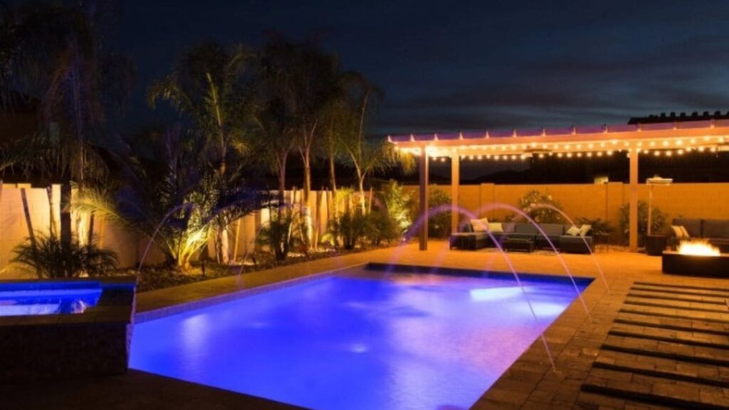 Backyard pool lit purple-blue at night with arcing water jets, a raised spa, string-lit pergola with seating, and uplit palm trees