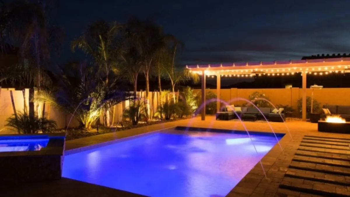 Backyard pool lit purple-blue at night with arcing water jets, a raised spa, string-lit pergola with seating, and uplit palm trees