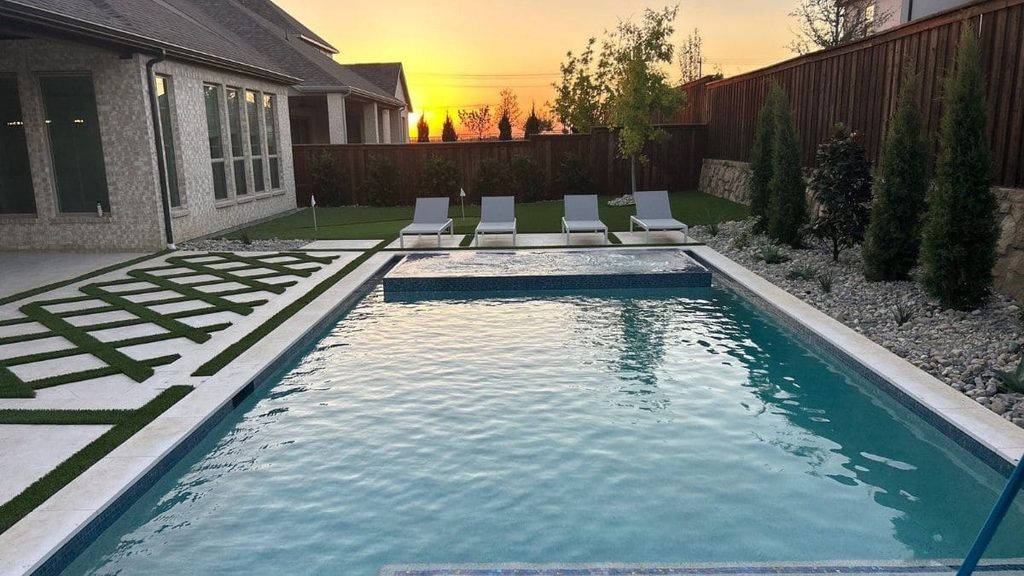 Rectangular backyard pool at sunset with a raised spa, white coping, geometric pavers set in artificial turf, four lounge chairs, and river rock landscaping