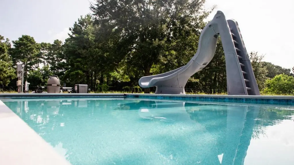 Backyard pool with clear turquoise water, a gray curved pool slide, visible pump and filter equipment, and tall trees in the background