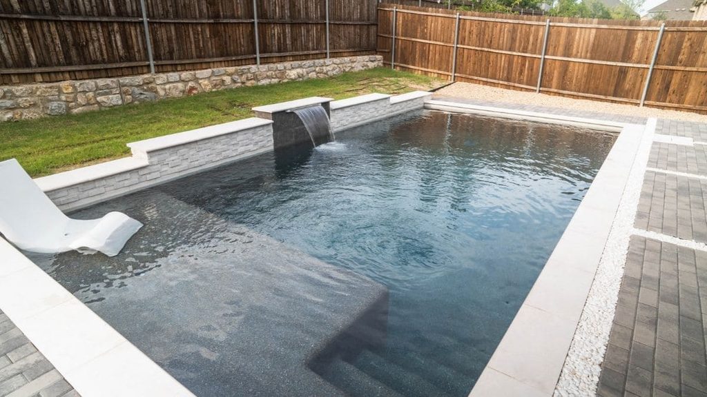 Backyard pool with a dark pebble finish, a stone spillway waterfall, a tanning ledge with an in-pool lounge chair, white coping, and a wood privacy fence