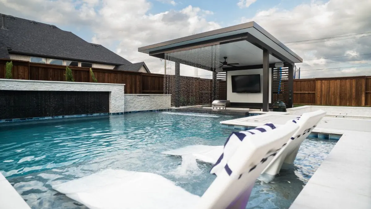 Backyard pool with in-pool lounge chairs, a stacked stone wall with a sheeting waterfall, and a covered outdoor kitchen with a TV and falling water curtain