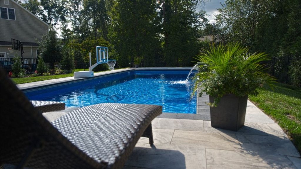 Backyard pool with a poolside basketball hoop, a small waterfall feature, a wicker lounge chair, a potted plant, and a stone paver patio