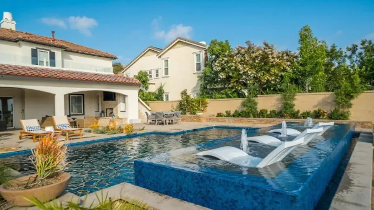 Backyard pool with a raised blue glass tile sun shelf, three in-pool lounge chairs with water bubblers, a covered patio with wood lounge chairs, and a stucco home with a tile roof
