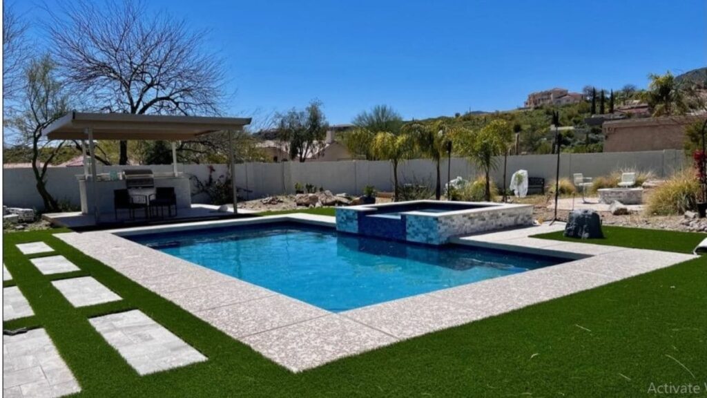 Backyard pool with a raised tile spa, textured concrete deck, artificial turf with stepping stones, and a covered outdoor kitchen in a desert hillside setting