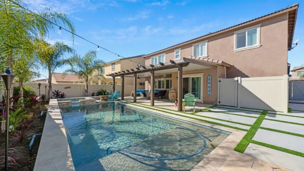 Backyard pool with a raised stone spa, travertine coping, covered patio with seating, string lights, palm trees, and concrete pavers set in artificial turf