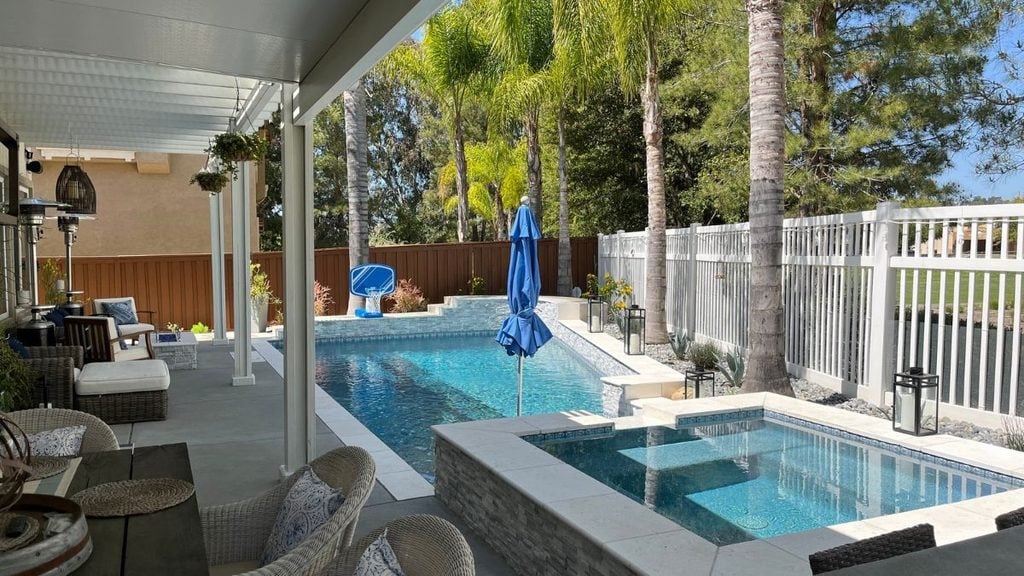 Backyard pool with a raised stacked-stone spa, a poolside basketball hoop, a covered patio with wicker seating and patio heaters, a blue umbrella, and tall palm trees