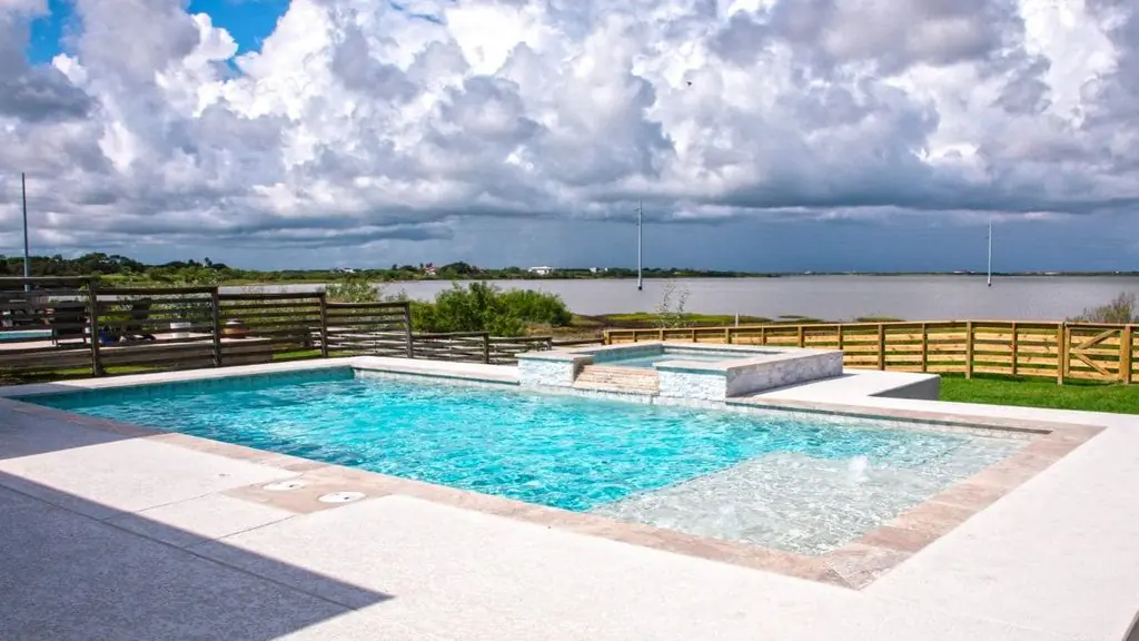 Rectangular backyard pool with a raised stone spa, tanning ledge with bubblers, light concrete deck, and a wood fence overlooking a bay under a cloudy sky