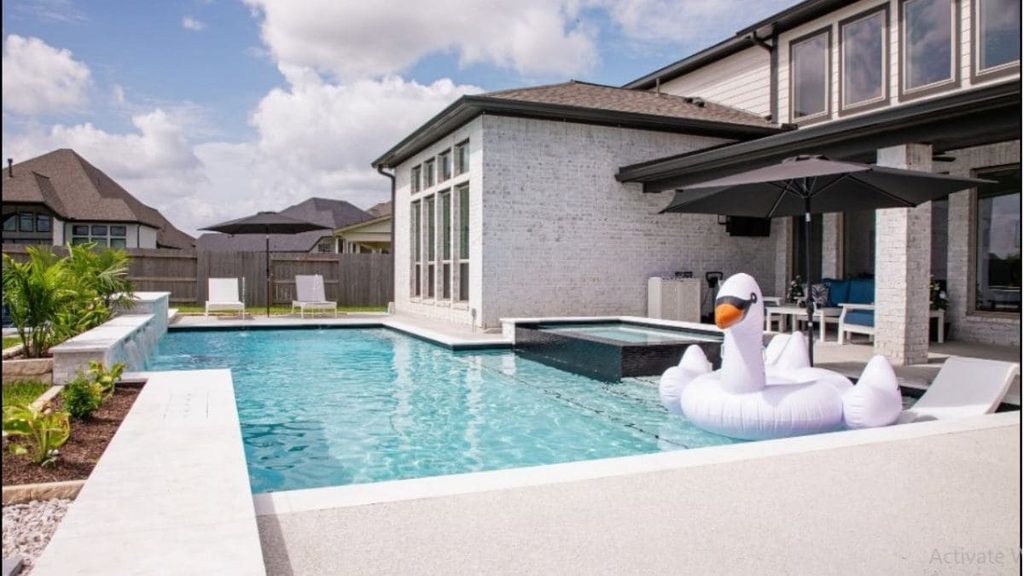 Backyard pool with a raised spa, stone water feature, in-pool lounge chair, and inflatable swan float beside a white brick home
