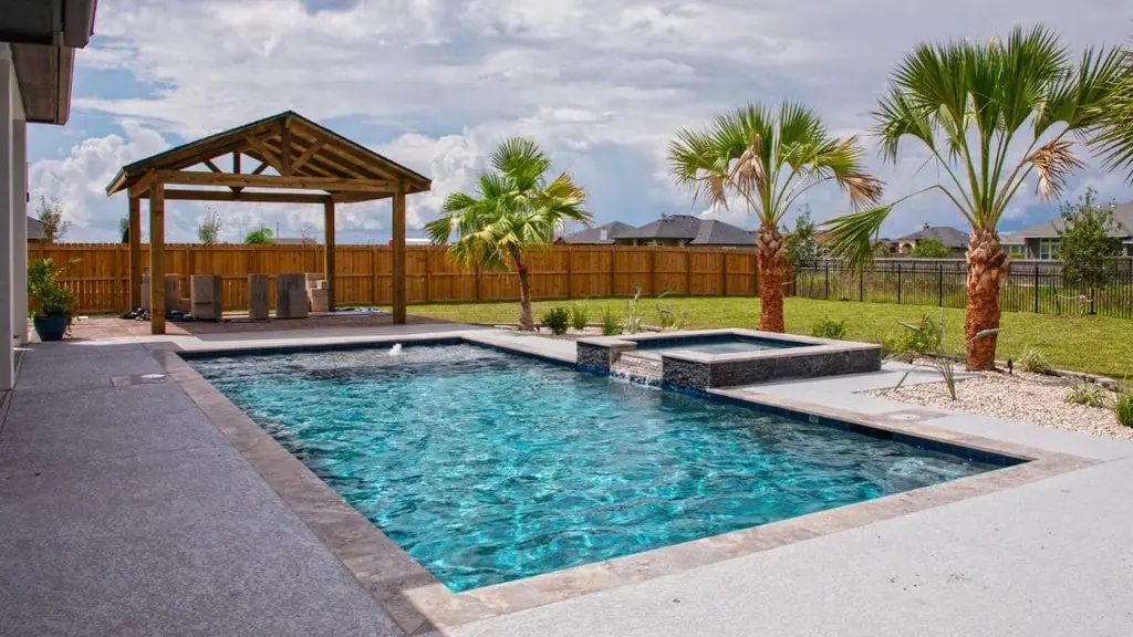 Rectangular backyard pool with a raised stone spa, textured concrete deck, a wood gazebo under construction, and palm trees along a grassy yard