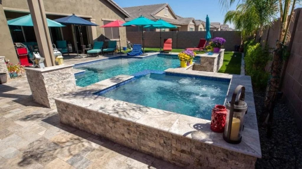 Backyard pool with a raised stacked-stone spa, bubbling jets, colorful patio umbrellas, lounge chairs, and a travertine paver patio