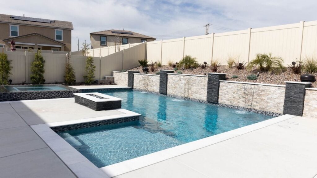 Rectangular backyard pool with a stacked stone retaining wall and sheeting water features, a raised spa, a square fire feature, white coping, and a light concrete deck