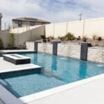 Rectangular backyard pool with a stacked stone retaining wall and sheeting water features, a raised spa, a square fire feature, white coping, and a light concrete deck