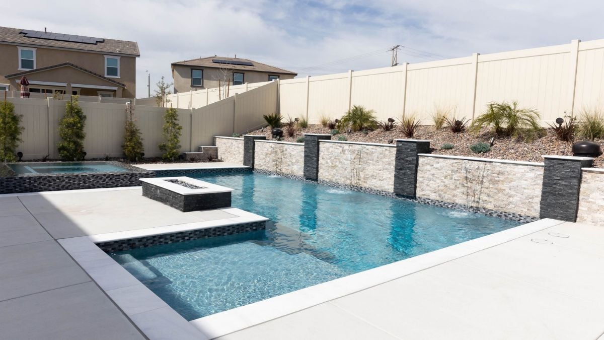 Rectangular backyard pool with a stacked stone retaining wall and sheeting water features, a raised spa, a square fire feature, white coping, and a light concrete deck