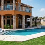 Backyard pool with a tanning ledge and in-pool lounge chairs, a raised stone spa, a travertine-style deck, and a covered patio with green Adirondack chairs beside a two-story stucco home