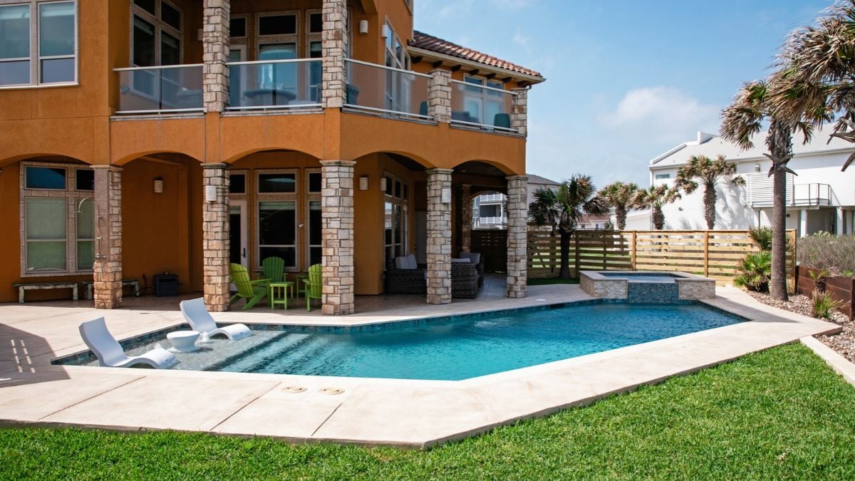 Backyard pool with a tanning ledge and in-pool lounge chairs, a raised stone spa, a travertine-style deck, and a covered patio with green Adirondack chairs beside a two-story stucco home