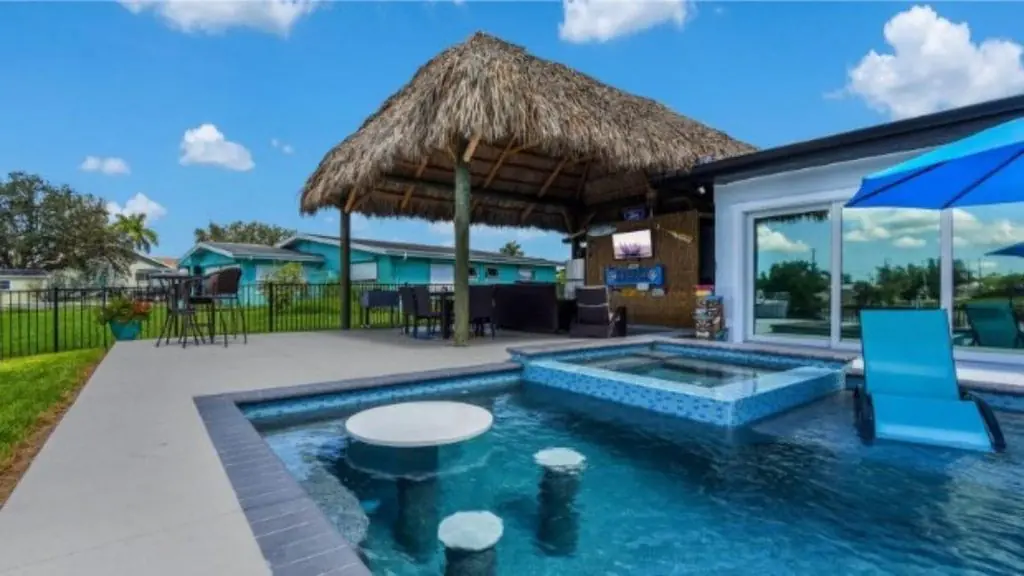 Backyard pool with a thatched tiki hut bar, in-pool swim-up table and stools, a blue tile raised spa, and a blue lounge chair and patio umbrella
