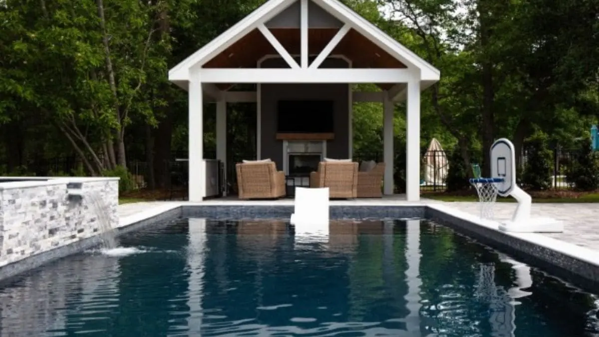 Backyard pool with dark water, a stacked stone waterfall wall, a poolside basketball hoop, and a white gabled pavilion with wicker seating and a fireplace