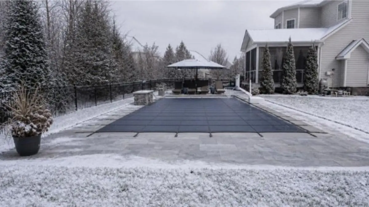 Backyard pool covered with a dark winter safety cover, surrounded by a stone patio, black metal fence, and a light dusting of snow