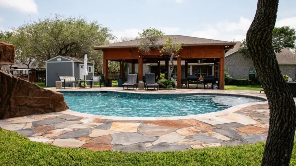 Freeform backyard pool with a flagstone deck, a rock feature on the left, lounge chairs, and a wood covered pavilion with outdoor kitchen seating