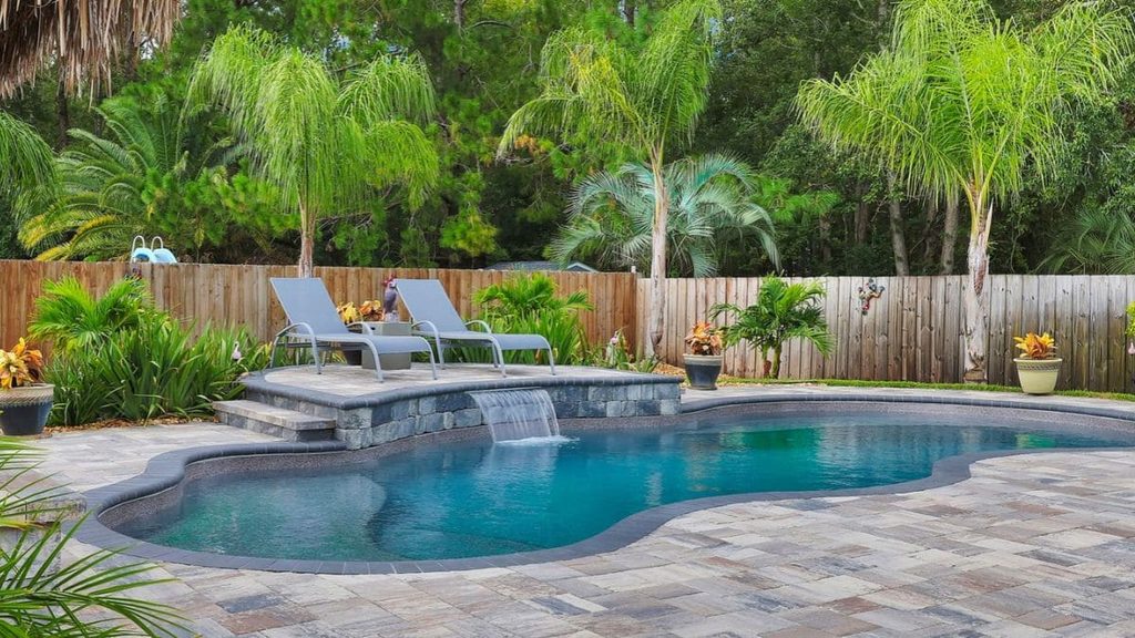 Freeform backyard pool with a stone waterfall feature, raised seating platform with two lounge chairs, paver deck, and palm trees