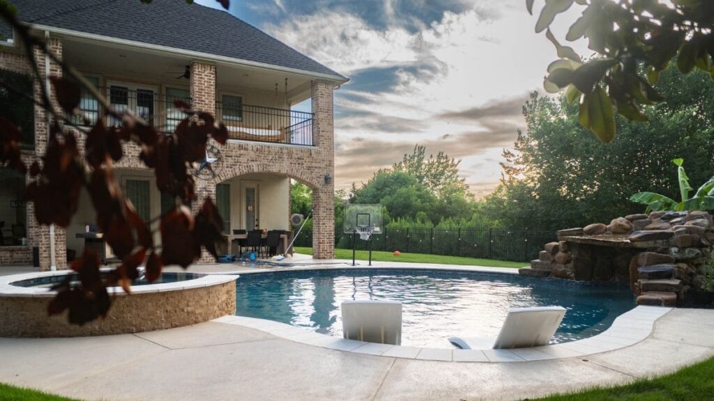 Freeform backyard pool with a rock grotto and waterfall, a raised stone spa, two in-pool lounge chairs, a poolside basketball hoop, and a brick home at sunset
