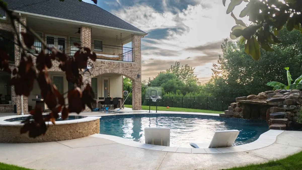 Freeform backyard pool with a rock grotto and waterfall, a raised stone spa, two in-pool lounge chairs, a poolside basketball hoop, and a brick home at sunset