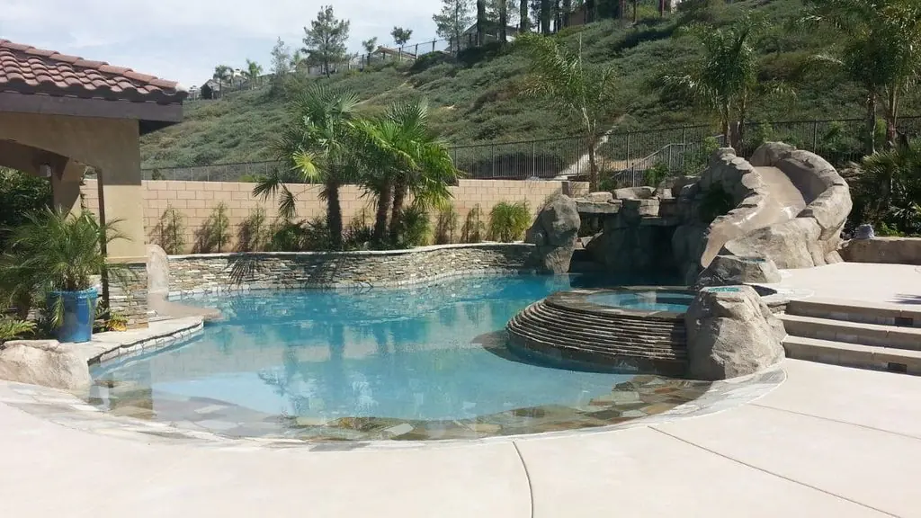 Freeform backyard pool with a rock slide and grotto, a raised stone spa with curved steps, a stacked stone wall, palm trees, and a hillside backdrop