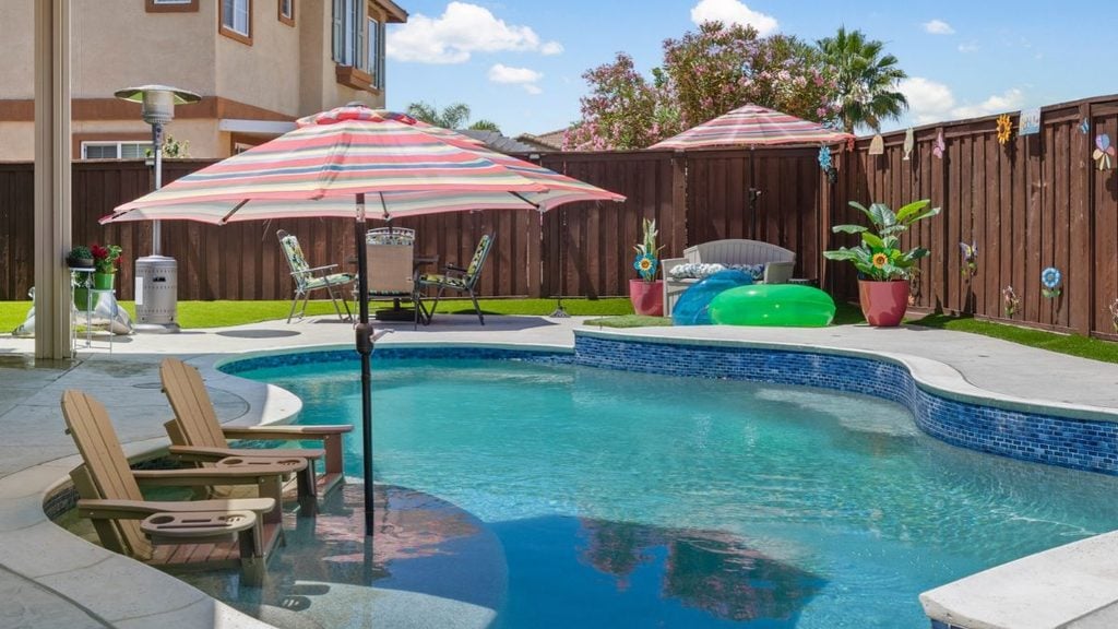 Freeform backyard pool with a blue tile wall, a tanning ledge with two Adirondack chairs, striped patio umbrellas, pool floats, and a wood privacy fence