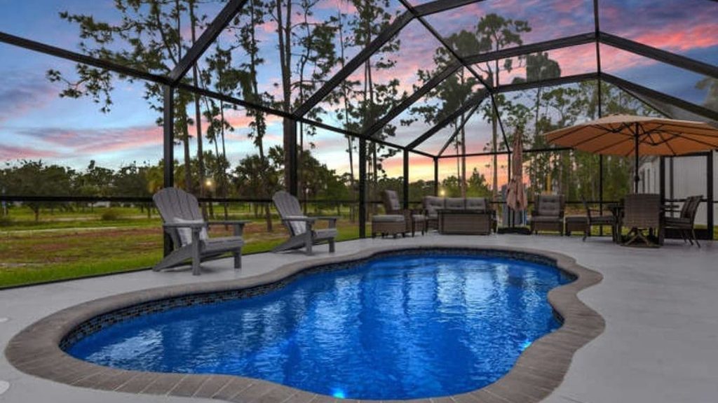 Freeform backyard pool inside a screened enclosure with a travertine deck, blue cushioned lounge chairs, a handrail, and tropical potted plants