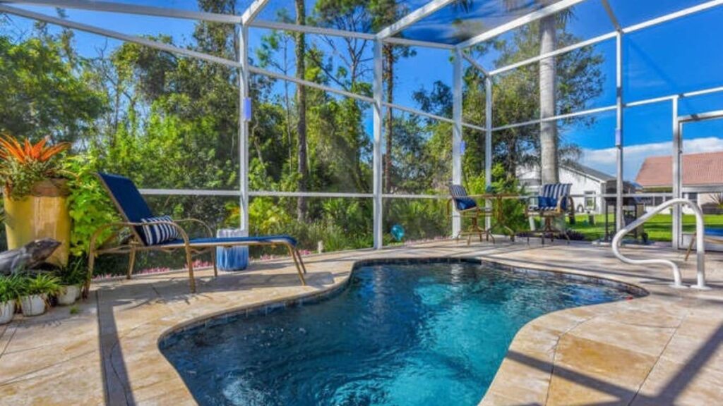 Freeform backyard pool inside a screened enclosure with a travertine deck, blue cushioned lounge chairs, a handrail, and tropical potted plants