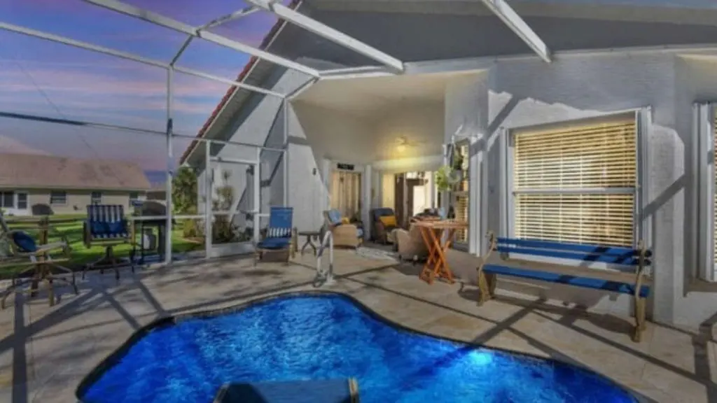 Freeform backyard pool with bright blue water inside a screened enclosure, with a travertine deck, a blue bench, and a covered patio seating area at dusk