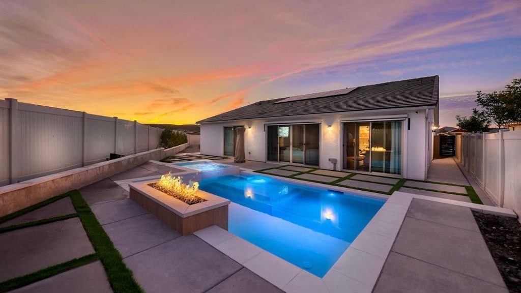 Modern backyard pool glowing blue at sunset with a linear fire feature, a raised spa, white coping, large concrete pavers set in artificial turf, and a stucco home
