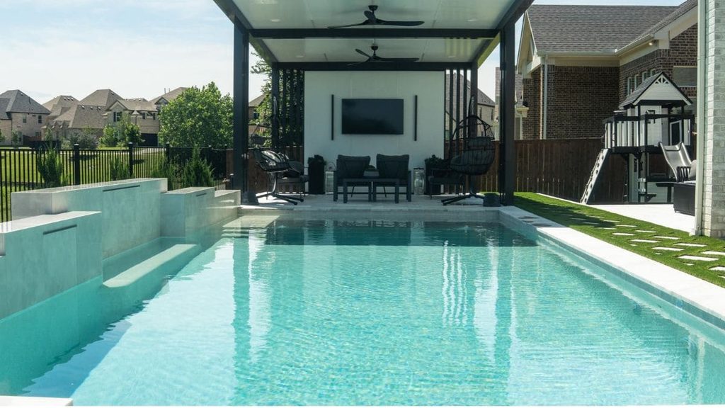 Modern backyard pool with light turquoise water, raised white water feature walls, a covered pavilion with ceiling fans, a TV, and black outdoor seating beside an artificial turf lawn