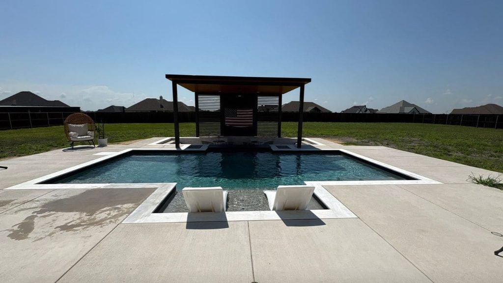 Modern backyard pool with two in-pool lounge chairs on a tanning ledge, a white coping edge, a covered pavilion with an American flag, and a large concrete deck