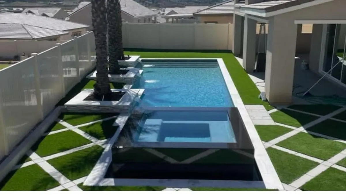Modern rectangular backyard pool with a raised glass-edge spa, stone water feature planters with palm trees, white coping, and diagonal pavers set in artificial turf