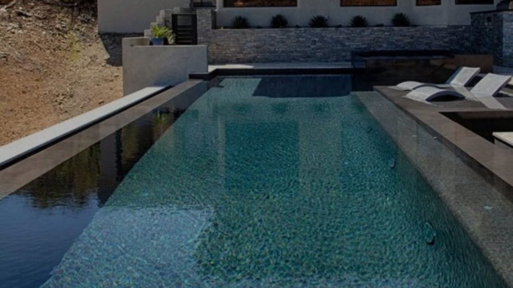 Modern infinity-edge pool with dark tile finish on a hillside property, with in-pool lounge chairs and a stacked stone wall