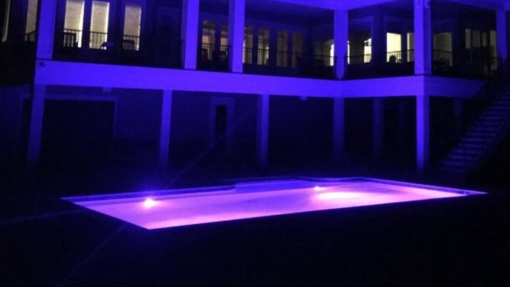 Modern rectangular swimming pool with purple LED lighting in a backyard beneath a two-story covered patio at night.